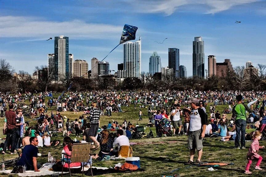 A large crowd spread across the great lawn at Zilker Park during the Kite Festival, with the Austin skyline beyond