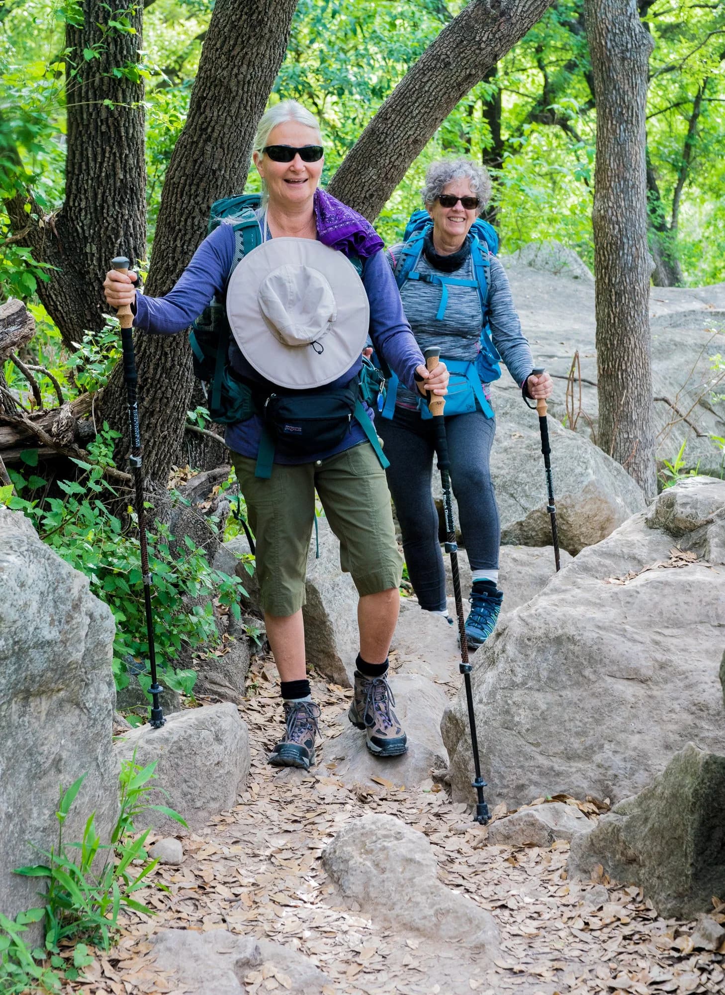 Two hikers with packs and trekking poles smiling on a rocky greenbelt trail