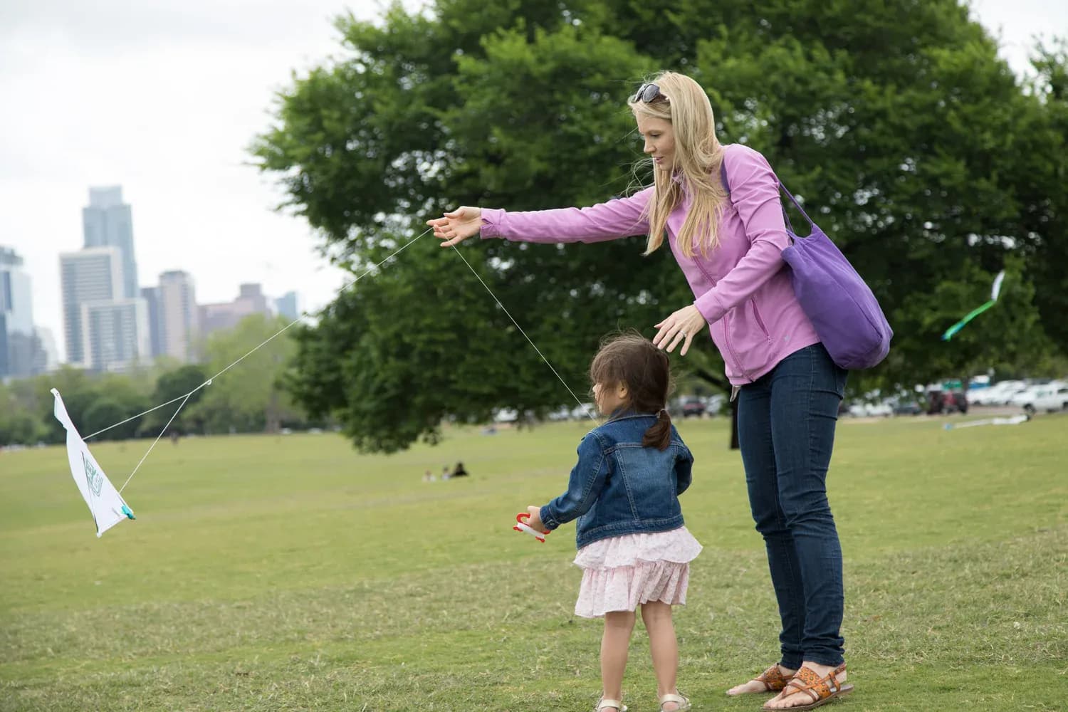 A parent helping a small child fly a kite on the lawn with the skyline behind