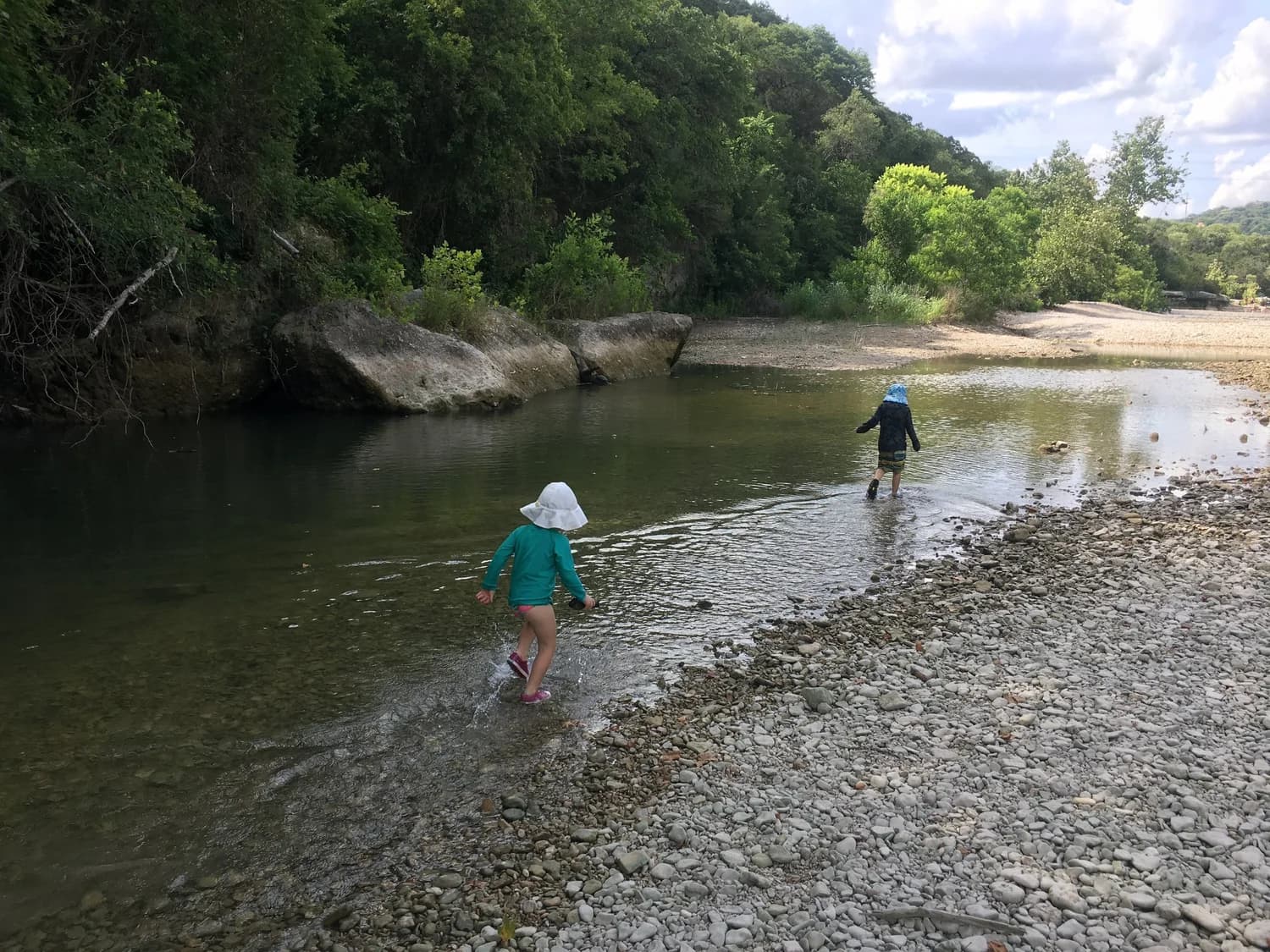 Two children in sun hats wading across the shallows of Barton Creek